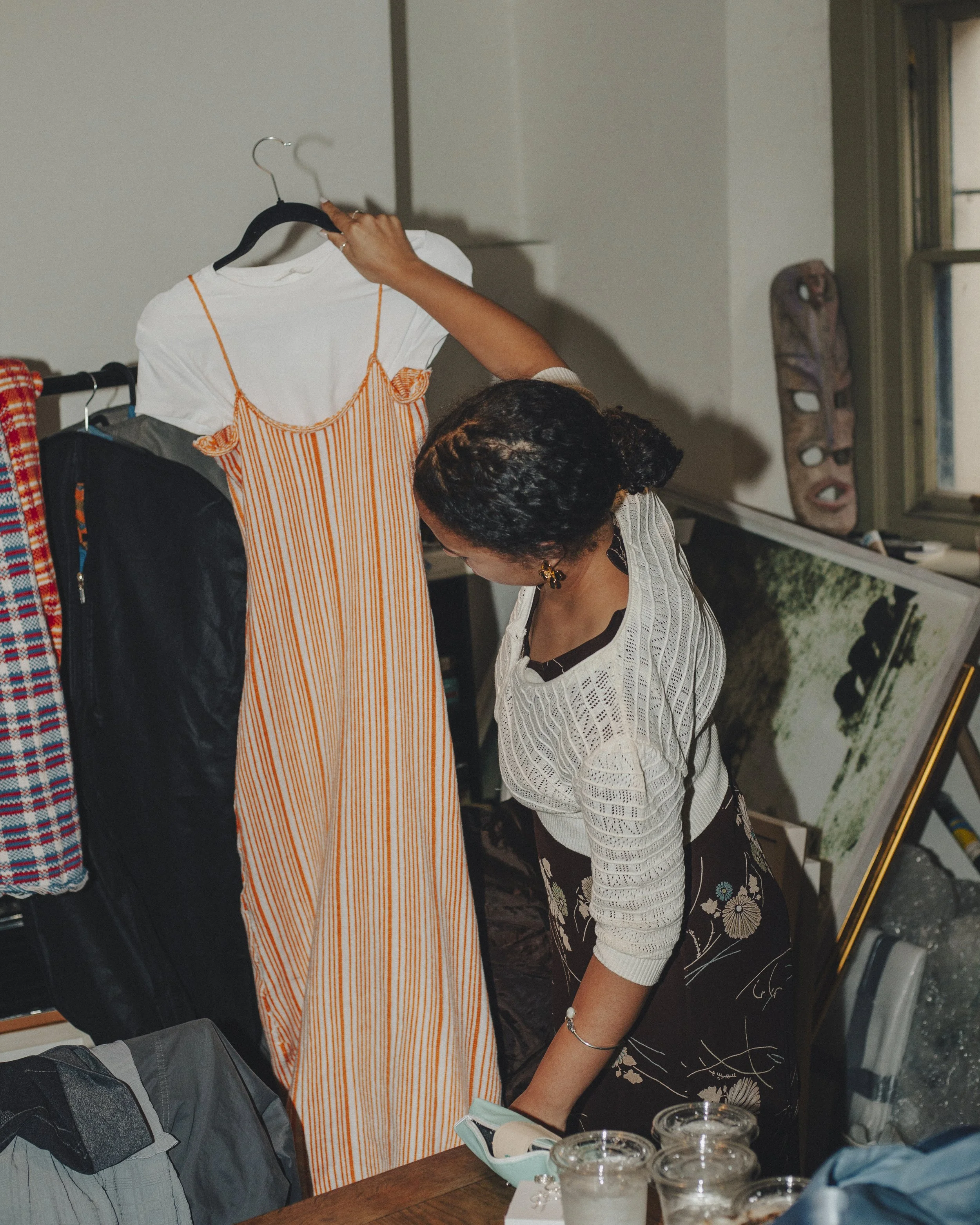 A woman with dark curly hair wearing a white knit sweater with black floral pattern, standing and helping another person. The person being helped is wearing a striped dress with orange and white vertical lines and is holding a white shirt. There are several glasses with drinks and a disposable face mask on the table, with a window and a tribal mask on the wall in the background.