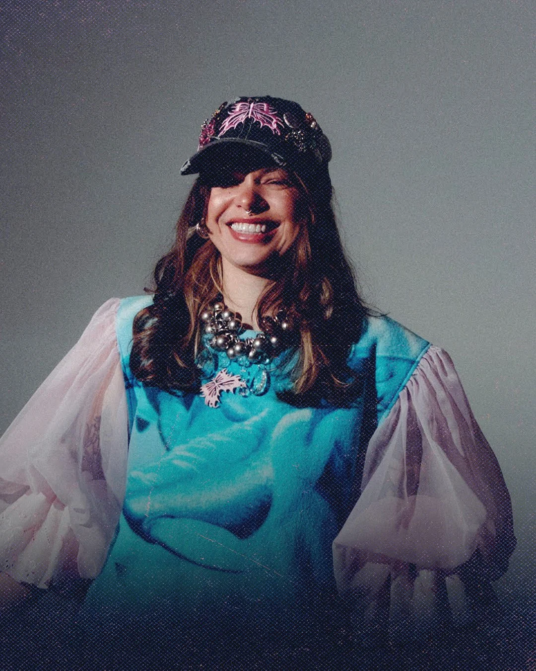 A woman smiling, wearing a black hat with pink embroidery, a blue top with sheer sleeves, a large pearl necklace, and hoop earrings.