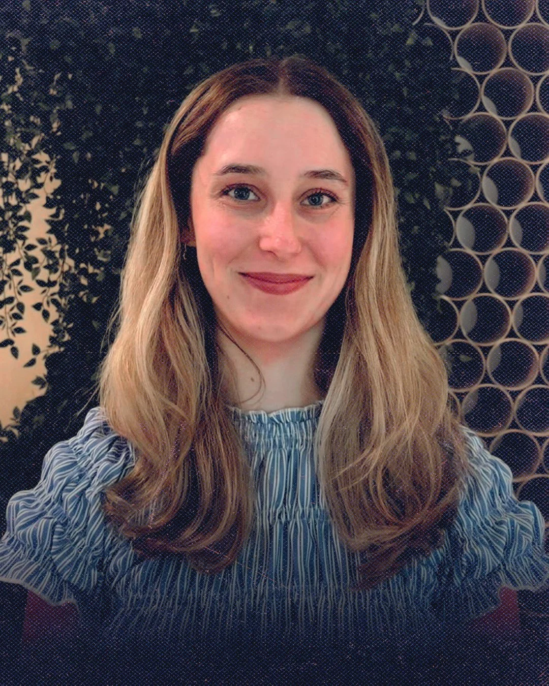 A woman with long, wavy light brown hair and blue eyes smiling at the camera. She is wearing a blue and white striped top. The background features a decorative black and gold circular patterned wall.