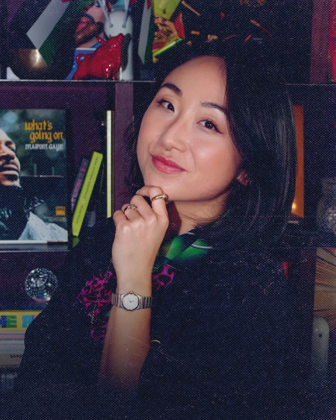 A woman with long black hair smiling and resting her chin on her hand, wearing a watch and rings, in front of a bookshelf with books and a TV screen.