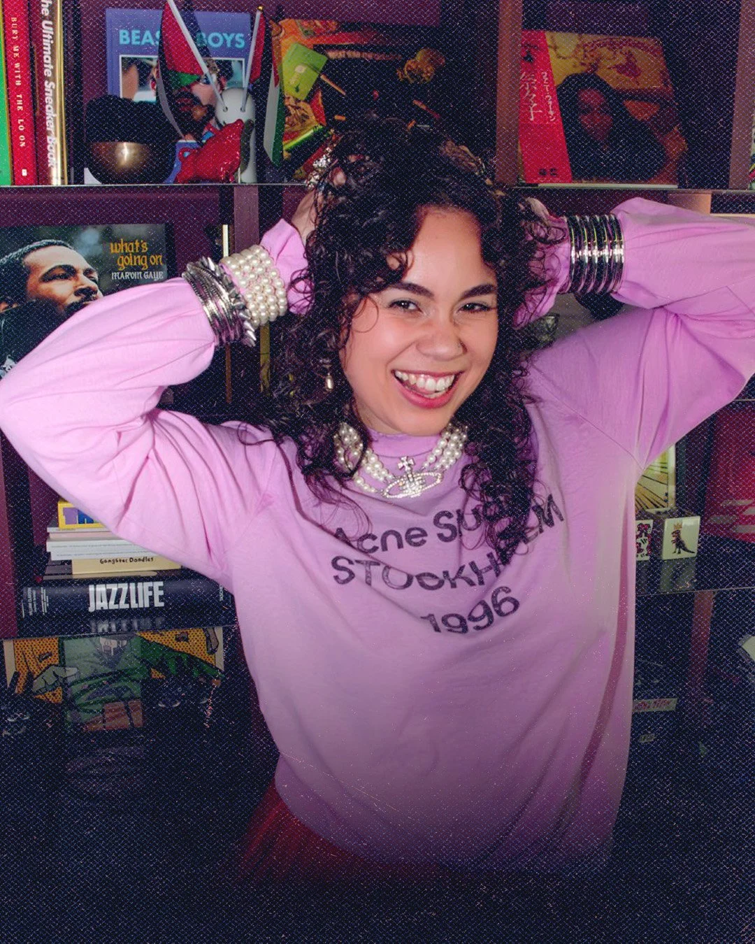A woman with curly dark hair, wearing a pink sweatshirt with black text, smiling and posing with hands behind her head, standing in front of a bookshelf filled with books and magazines.