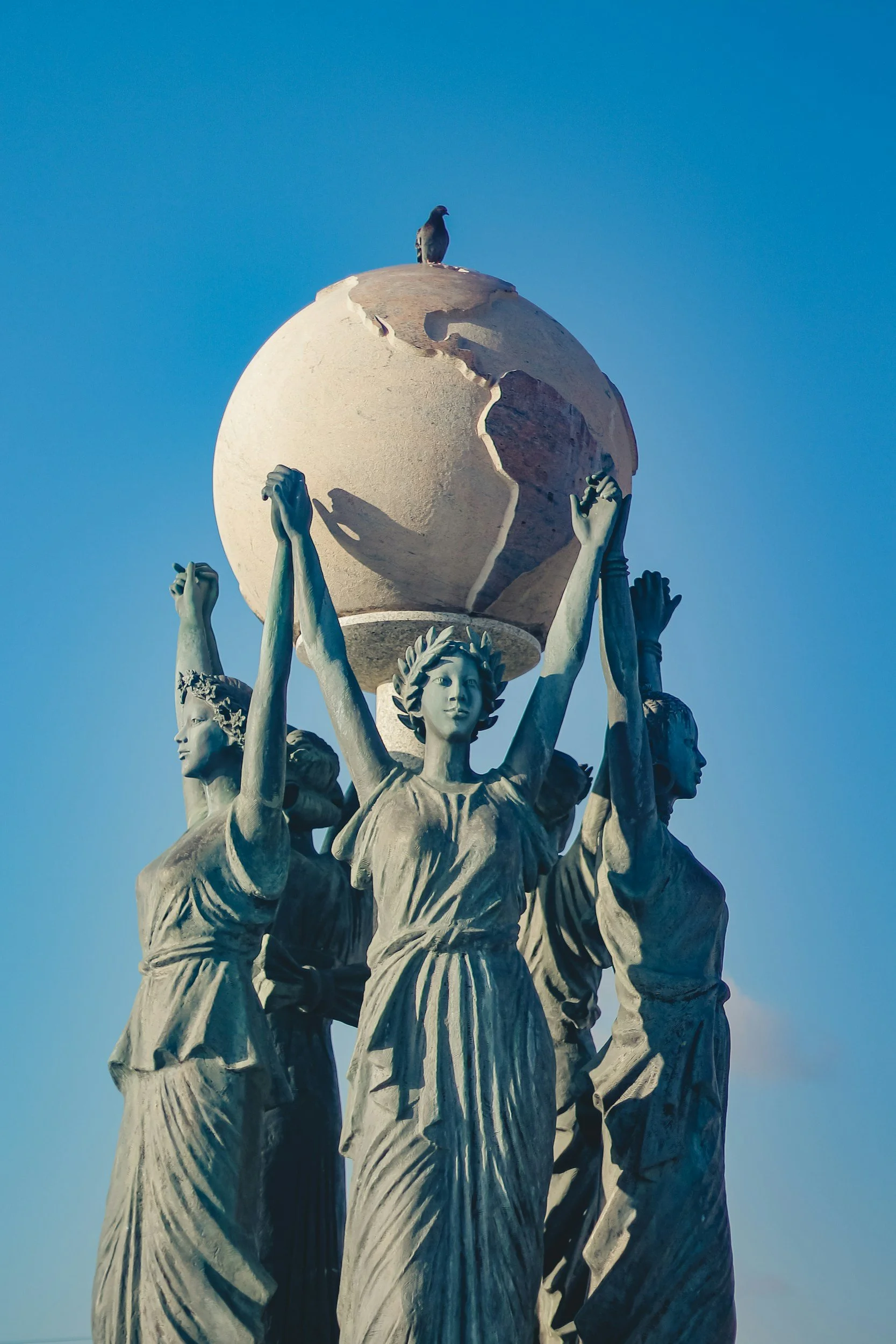 statue of women standing in a circle facing out while holding hands with their hands up in the air and the world in the middle.
