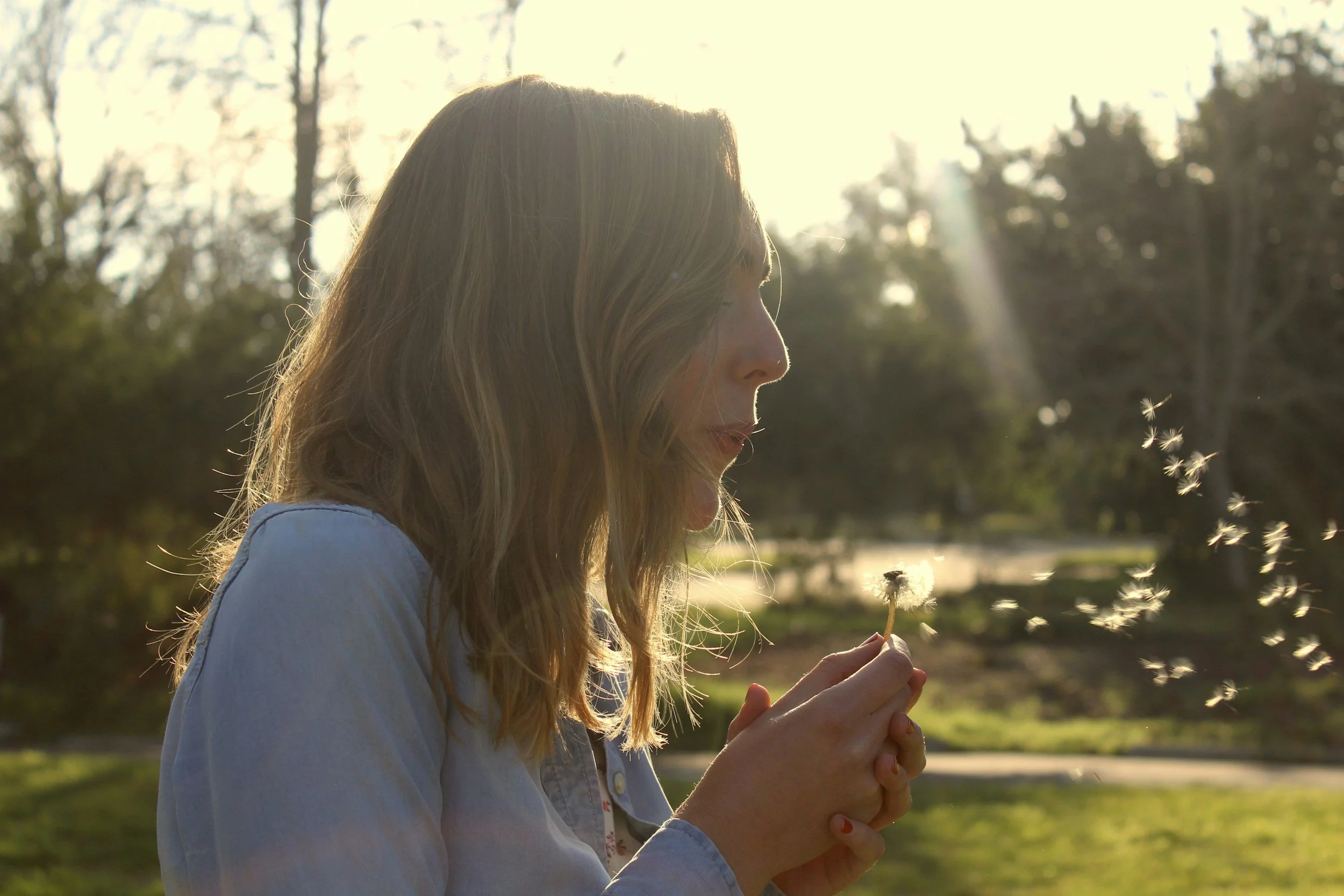 A side profile of a woman outside blowing on a dandelion flower.