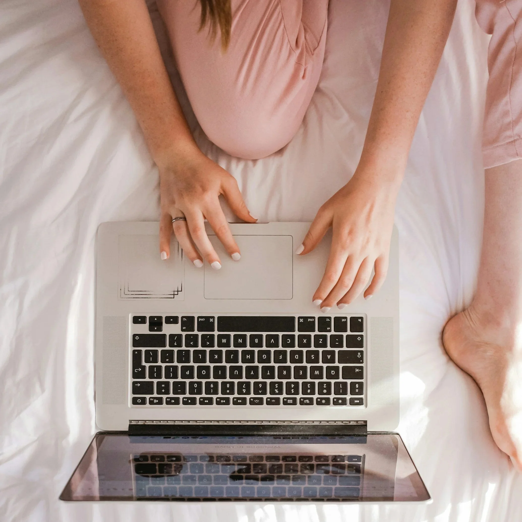 woman sitting on bed with two hands and a foot visible. Her hands are on her laptop computer.