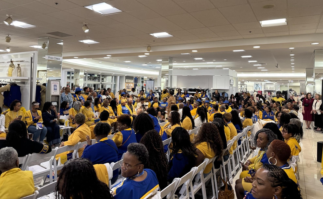 A large group of people gathered in a shopping mall, many wearing yellow and blue clothing, seated in white chairs, attending an event or presentation.