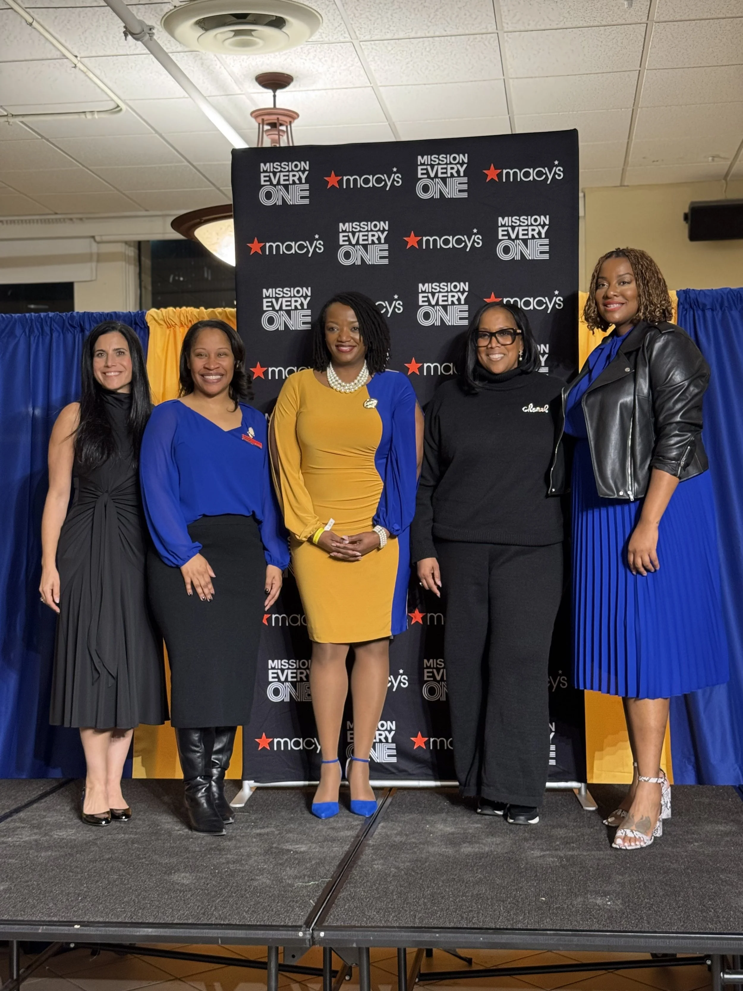 Five women standing on a stage in front of a backdrop with the Macy's logo and the slogan 'Mission Every One'. They are dressed in formal attire, smiling, and posing for a photo.