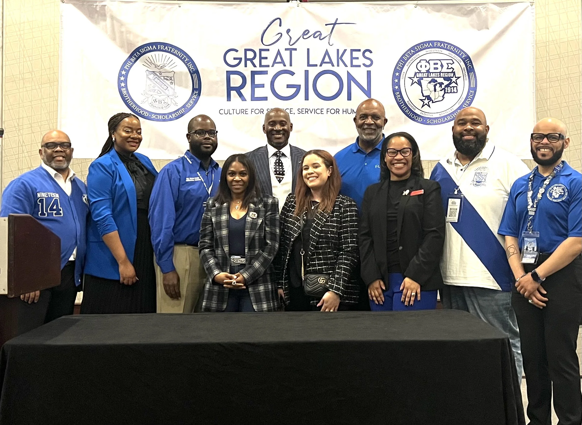 A group of men and women posing in front of a banner that reads "Great Lakes Region" with the Phi Beta Sigma Fraternity emblem on each side. The people are smiling and dressed in formal and business casual attire, with some wearing fraternity shirts and badges.