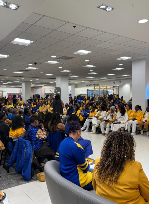 Group of people gathered in a store, many wearing matching yellow and blue clothing, seated in a semi-circle, listening to a speaker or presenter.