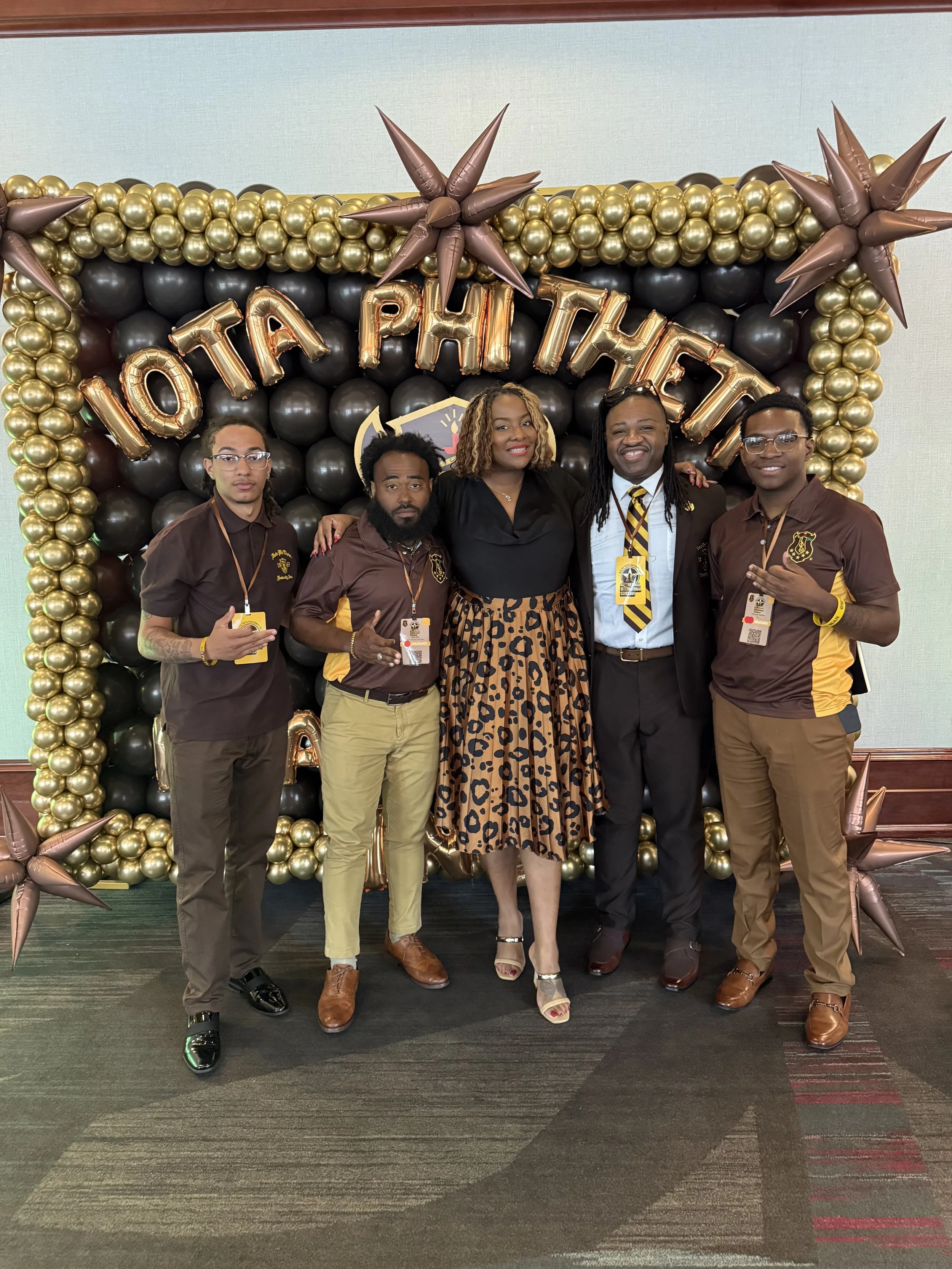 Group of five people standing in front of a balloon backdrop with gold and black balloons and metallic stars, celebrating a Phi Theta Kappa event, with the word 'CONGRATULATIONS' spelled out in large balloons above them.