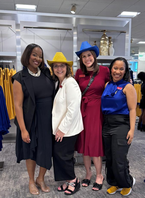 Four women standing together inside a store, smiling, wearing colorful hats and business casual attire.