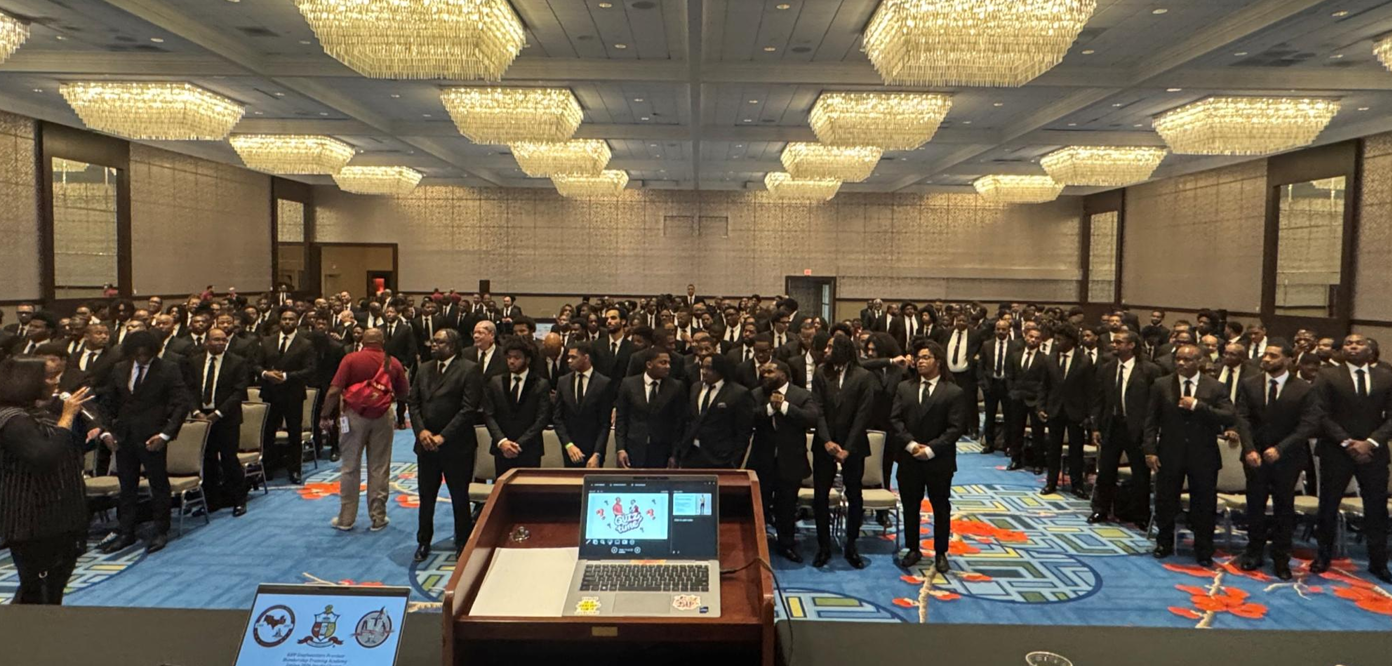 Large conference room filled with many men dressed in black suits and ties standing and listening to a speaker. The room has chandeliers and a blue carpet with orange and yellow patterns.