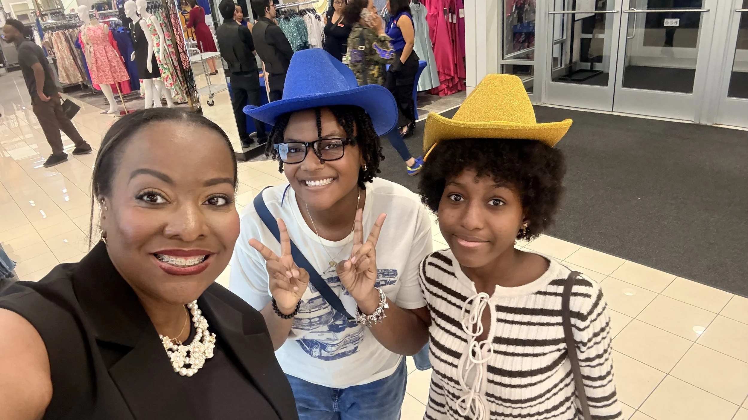 Three women and a girl taking a selfie inside a shopping mall, with clothing displays and people shopping in the background.