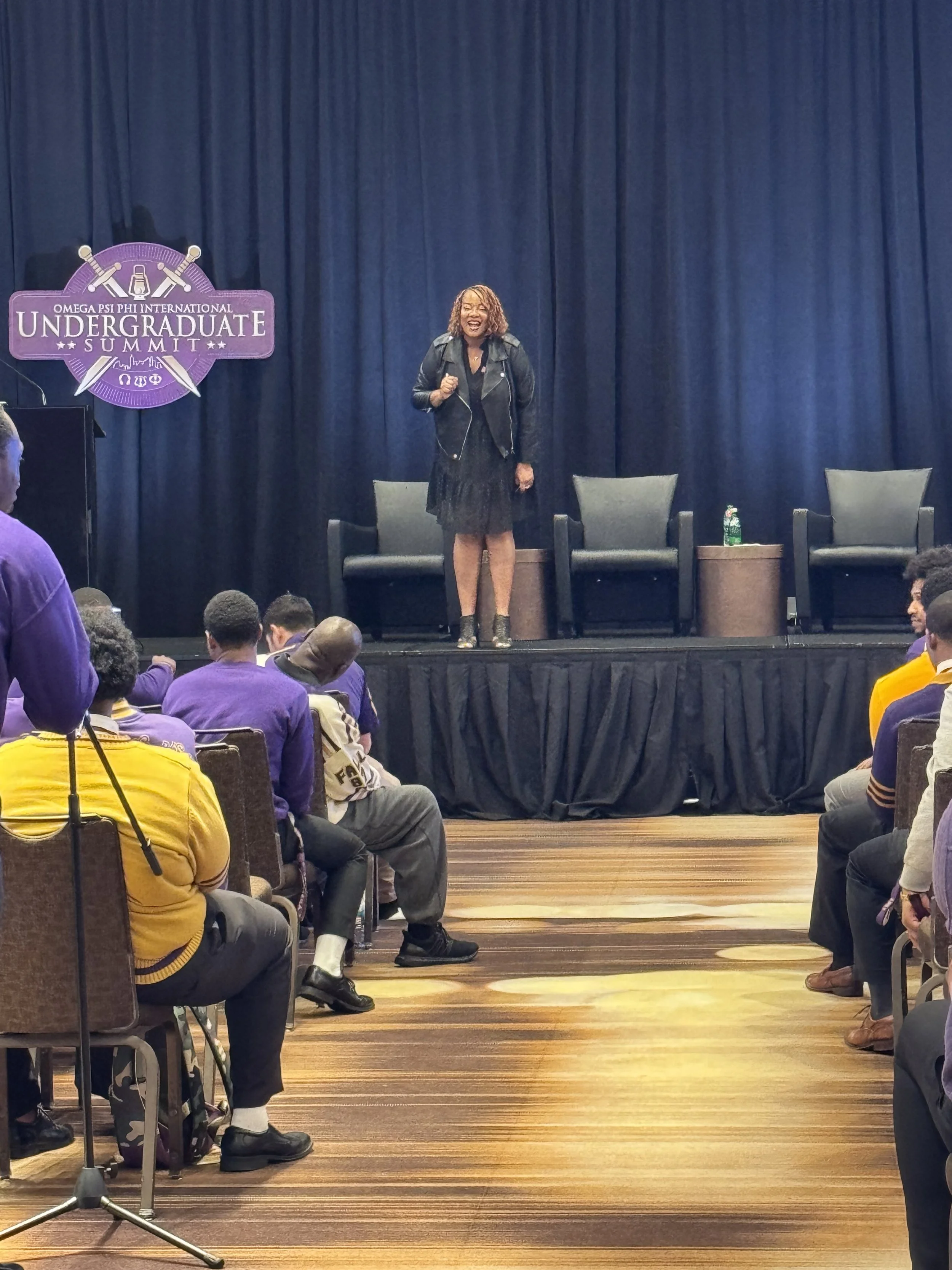 A woman standing on a stage with a blue curtain background, speaking or presenting to an audience at the Omega Psi Phi International Undergraduate Summit. The audience members are seated, and some are wearing purple or yellow clothing. There are chairs on the stage and a table with a water bottle.