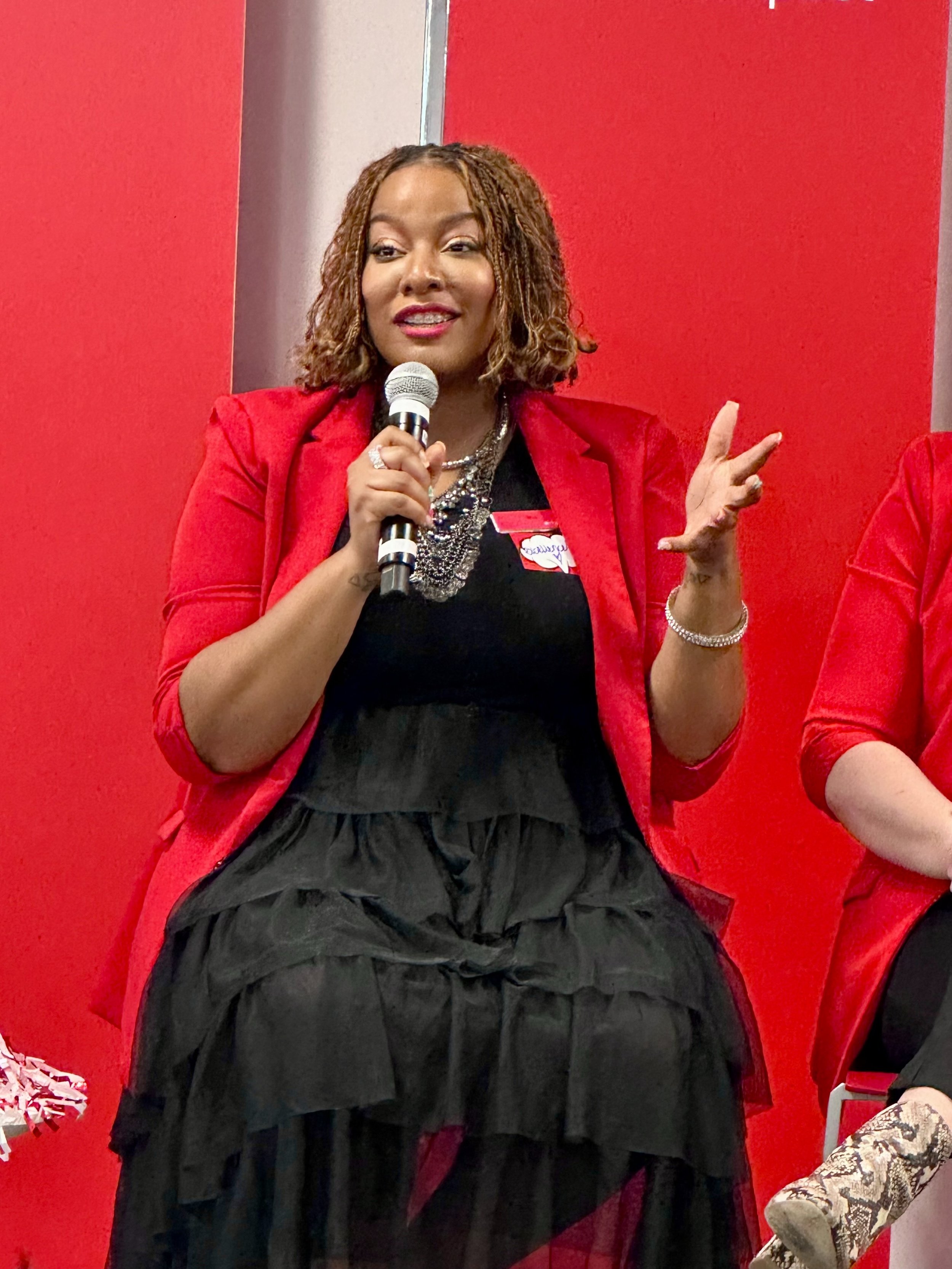 A woman with brown curly hair wearing a red blazer, holding a microphone, and gesturing with her right hand, standing in front of a red wall.