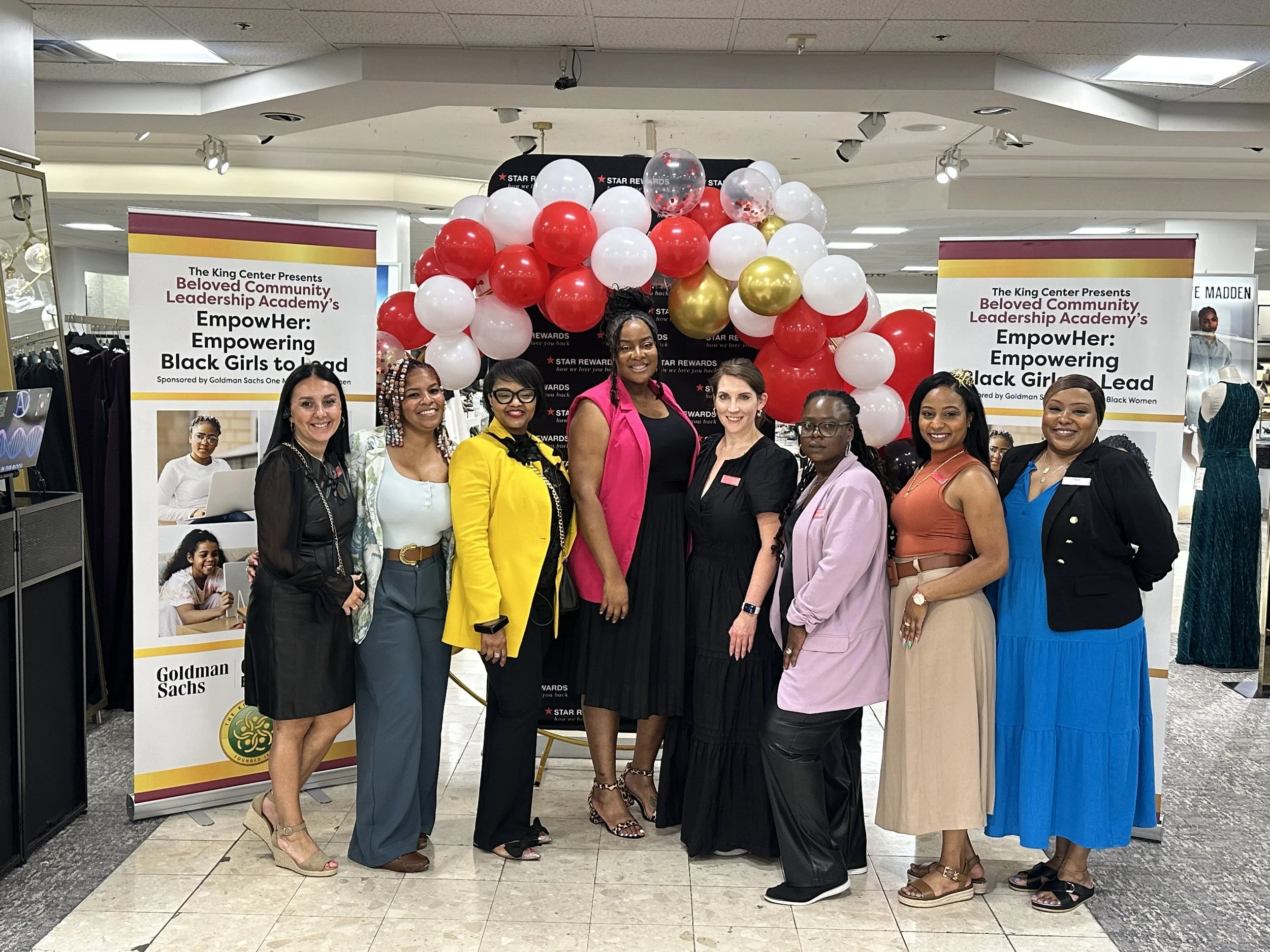 A group of eight women standing together in front of a balloon display and banners for the Beloved Community Leadership Academy event at The King Center. The women are smiling and dressed in various stylish outfits.