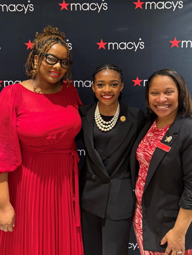 Three women standing together at Macy's event, smiling. The woman on the left is wearing a red dress and glasses, the woman in the middle is in a black blazer with a pearl necklace, and the woman on the right is in a black blazer with a red and white patterned shirt.