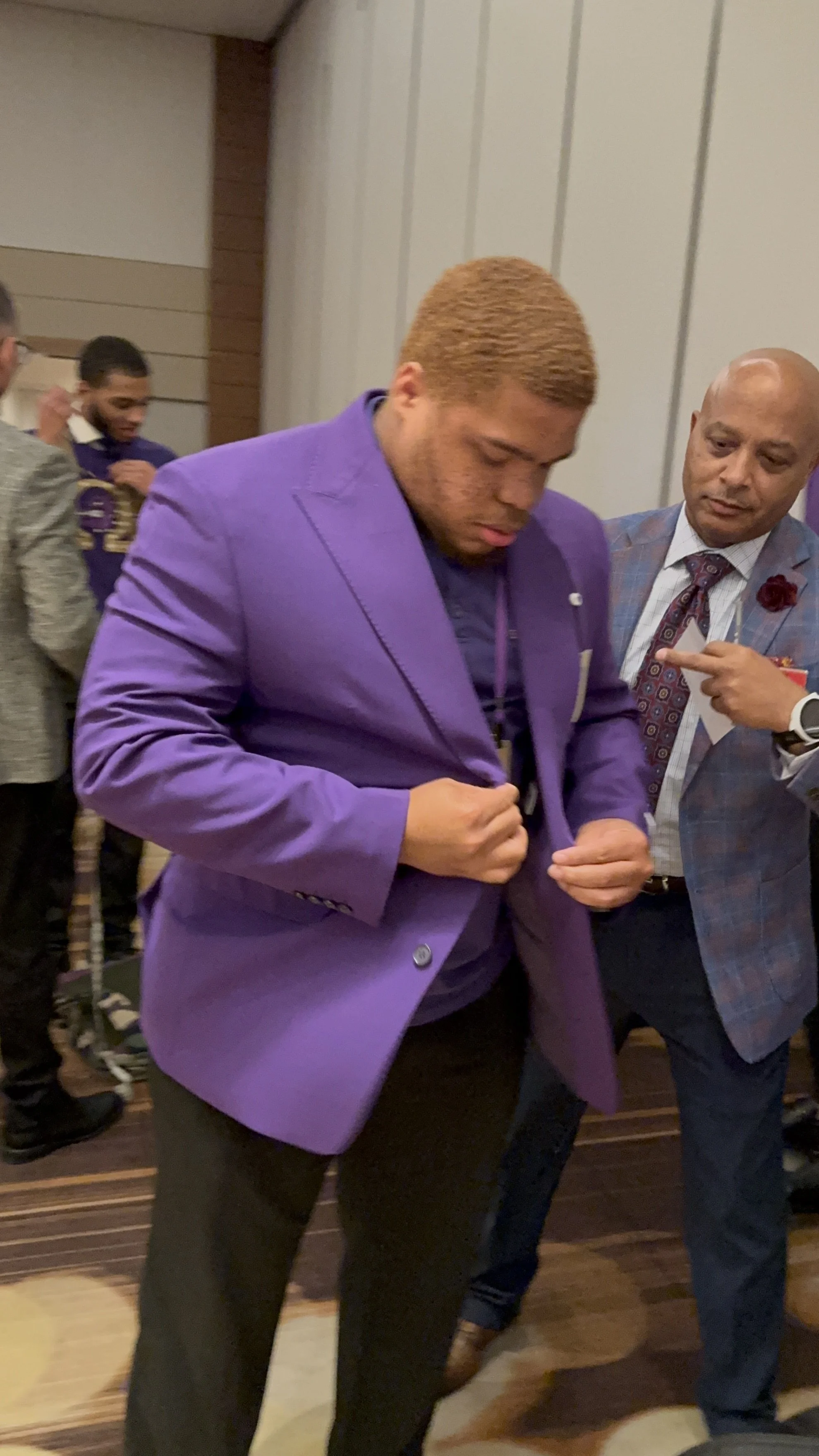 Man with blond hair wearing a purple suit adjusting his jacket at an event, with another man in a checked suit pointing at him, and others in the background.