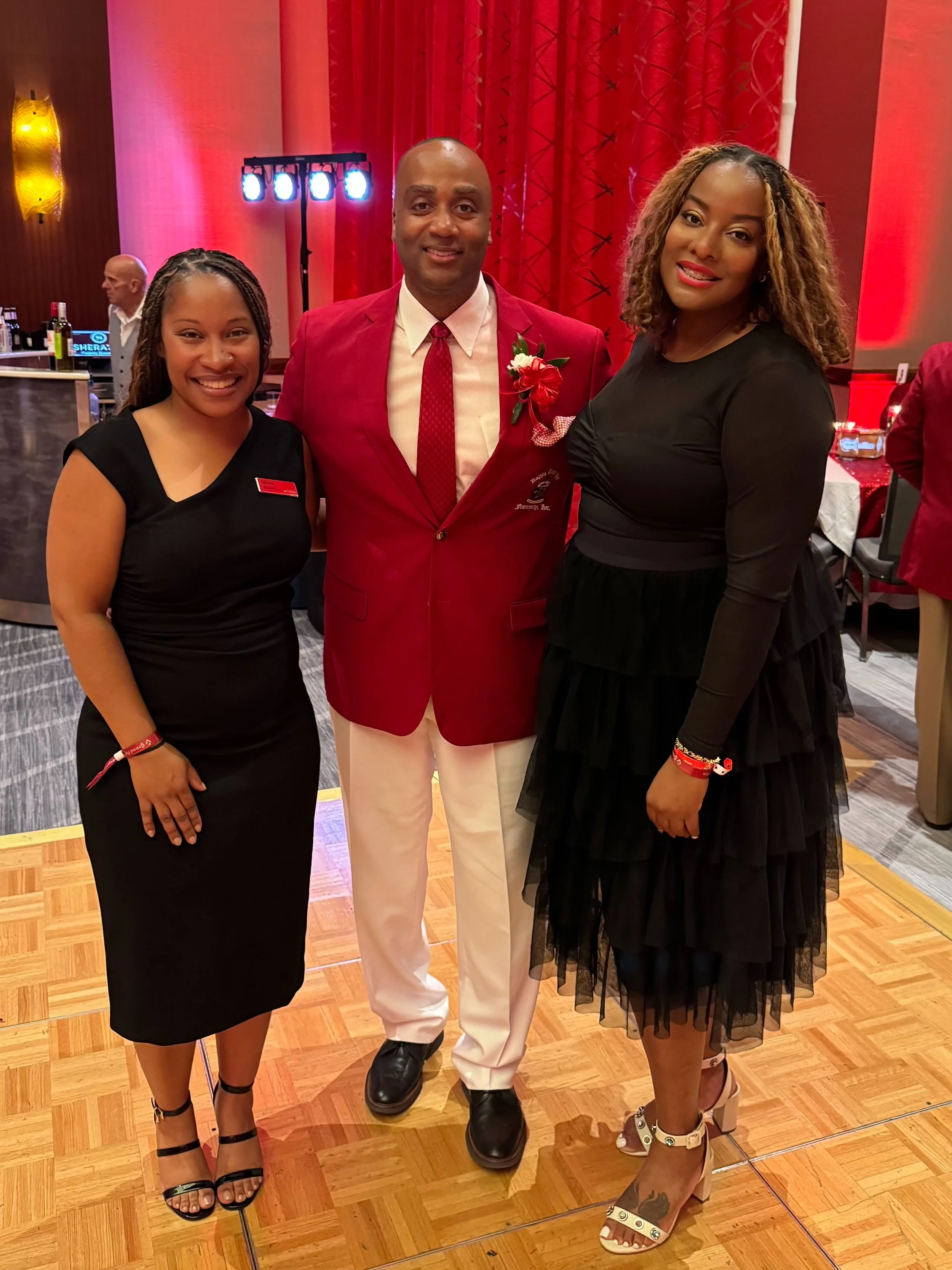 Three people posing together at an event: a woman in a black dress on the left, a man in a red suit in the middle, and a woman in a black dress with a tulle skirt on the right, in a venue with red curtains and wooden flooring.