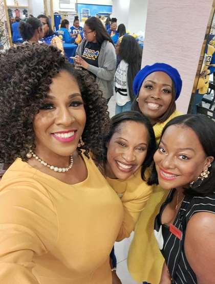 Four women smiling for a selfie at an indoor event, with people in the background.