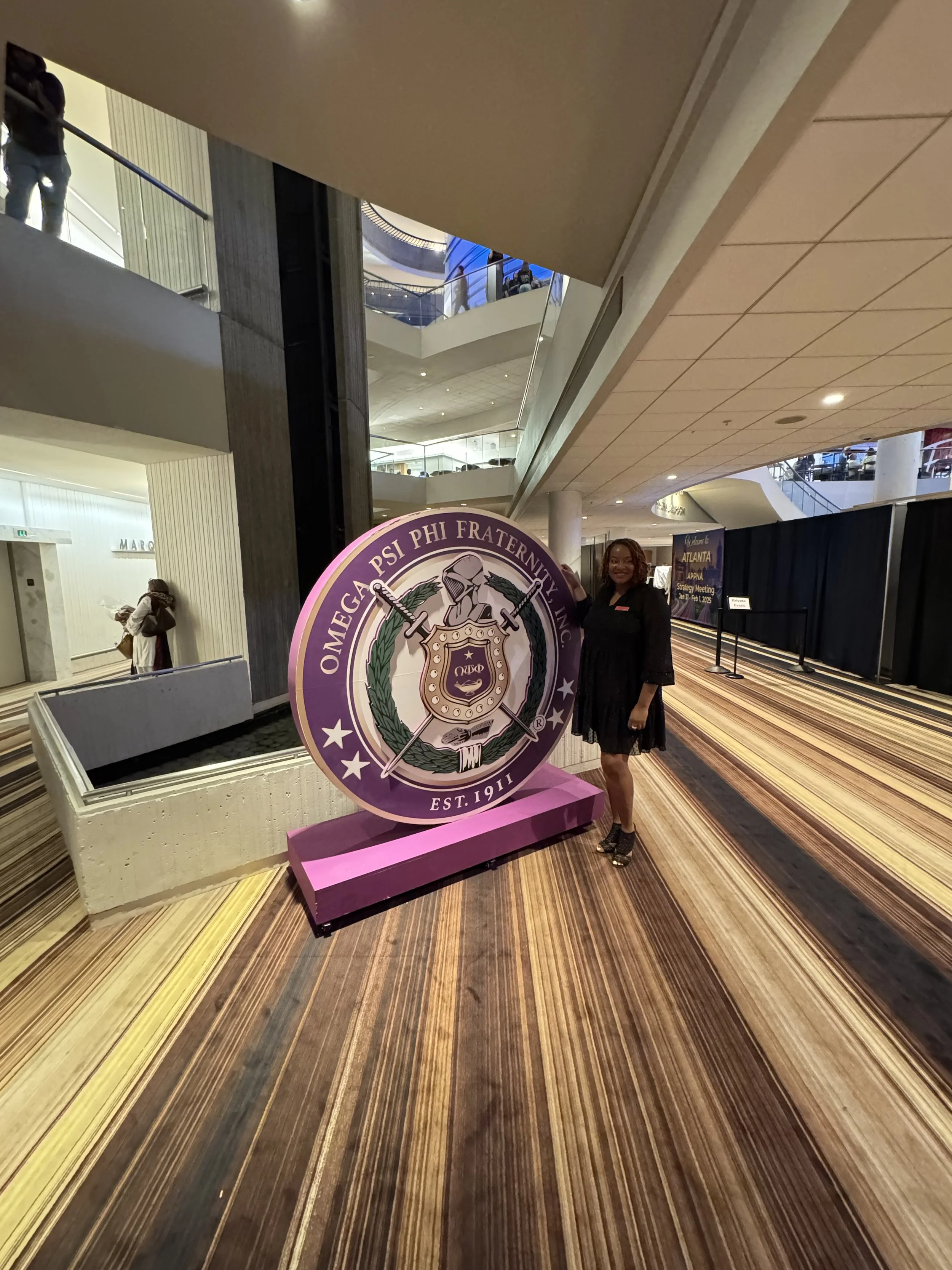 A woman in a black dress standing next to a large circular sign for Omega Psi Phi Fraternity Inc. at a convention center with multiple floors and escalators in the background.