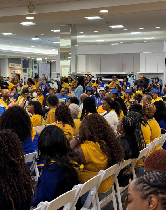 A large group of people wearing yellow and blue uniforms gathered in a retail store for a meeting or event, with some seated on white folding chairs and others standing.
