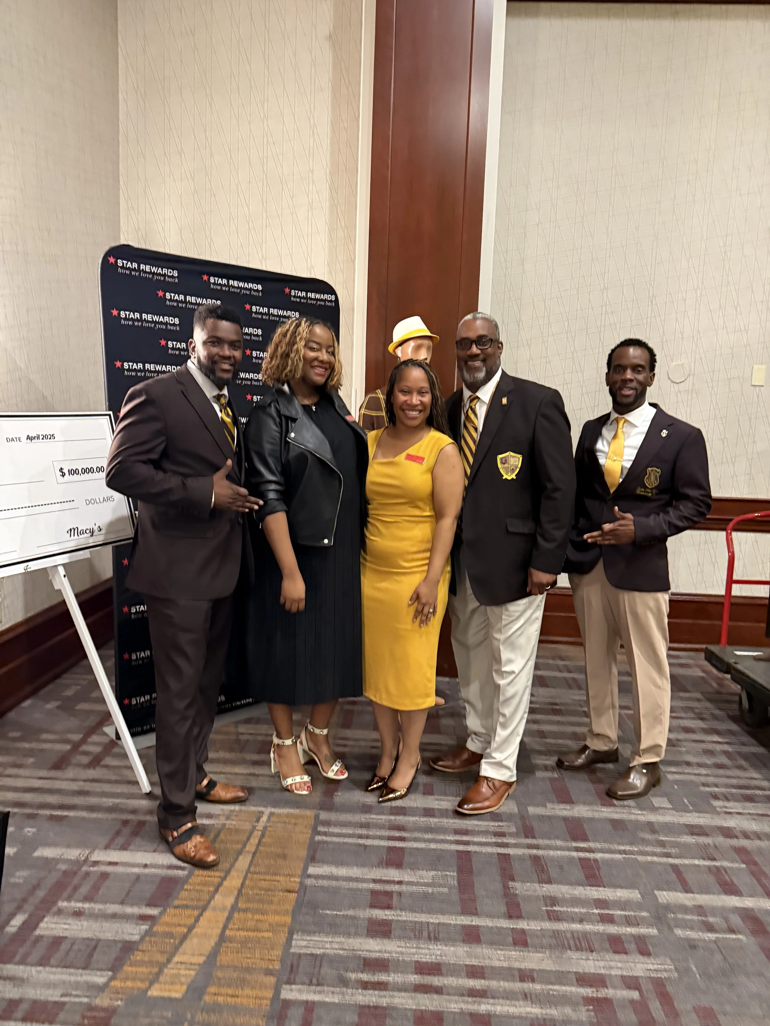 Group of six people dressed in formal and semi-formal attire standing in a line at an indoor event. They are smiling and posing for the photo, with a backdrop that reads 'Star Rewards' and has a large check referencing $100,000.