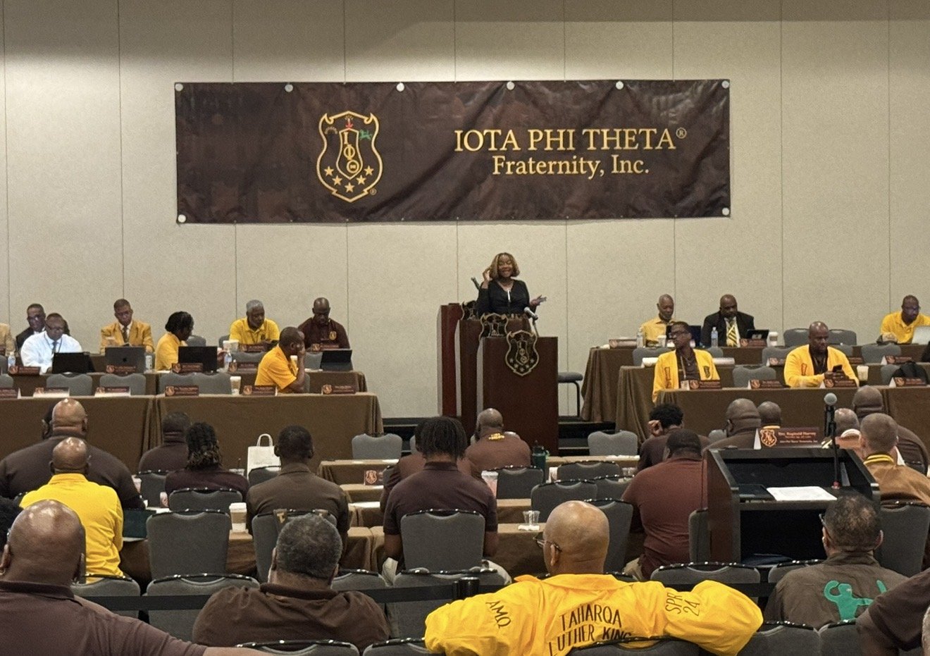 A woman speaking at a podium during a conference for Iota Phi Theta Fraternity, Inc., with attendees in yellow and brown shirts seated in front, some wearing fraternity regalia, in a large meeting hall.