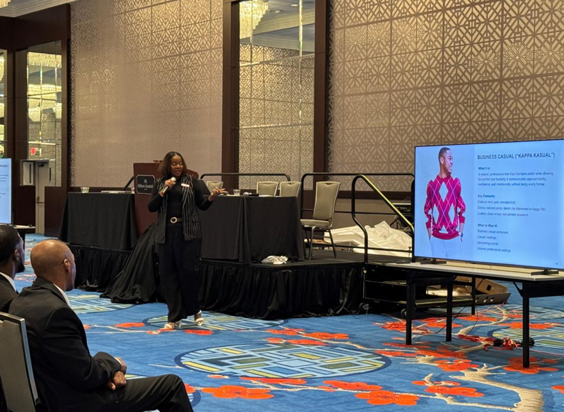 A woman giving a presentation in a conference room, standing near a large screen displaying a slide about business casual attire, with two seated attendees watching her.