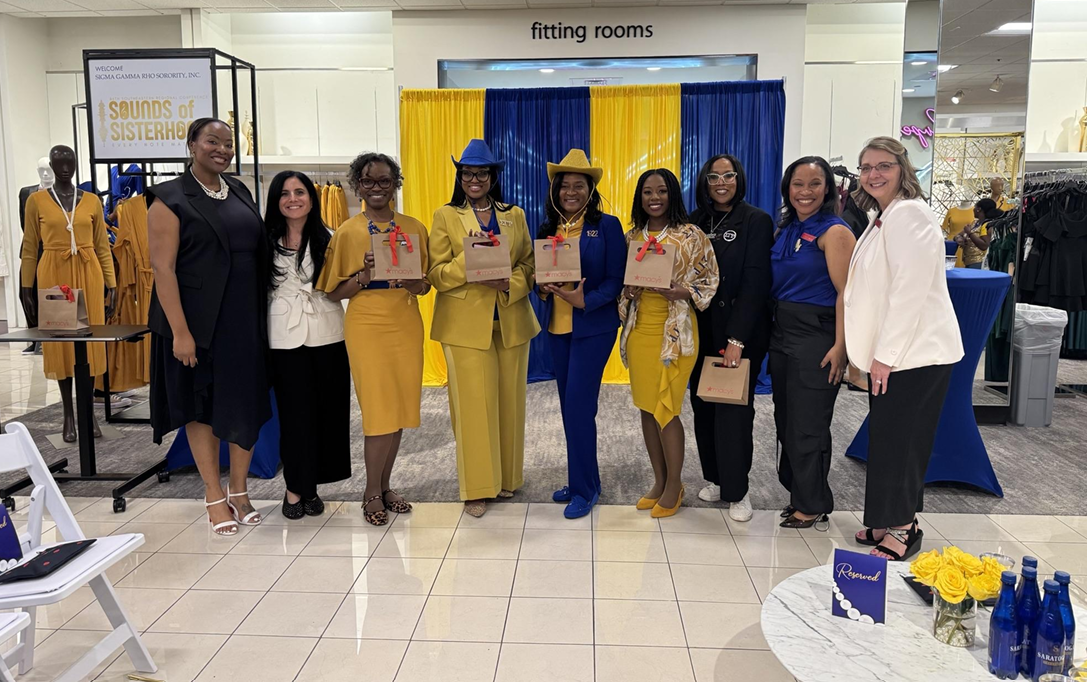 Group of women standing in front of a fitting room area at a shopping mall, holding gift bags, with a blue and yellow curtain backdrop behind them.