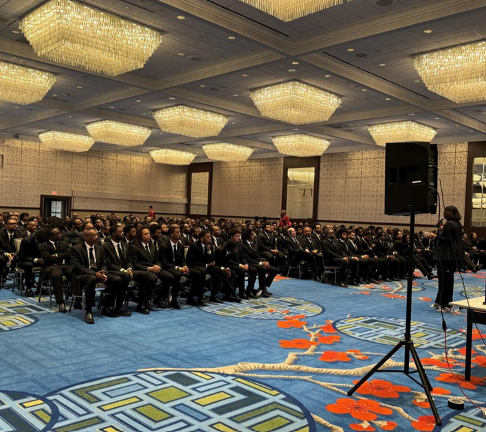 Large conference room filled with formally dressed people seated in rows, listening to a speaker at a podium, under ornate chandeliers.