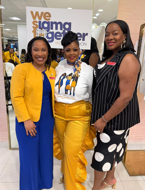 Three women smiling and standing together at an event, with a sign that reads 'We Sigma Together' in the background. They are dressed in colorful outfits, with one in blue and yellow, one in yellow pants and a white top, and one in a black and white polka dot dress.