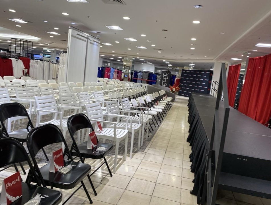 Empty chairs arranged in rows for an event setup inside a retail store, with clothing racks and red curtains in the background.