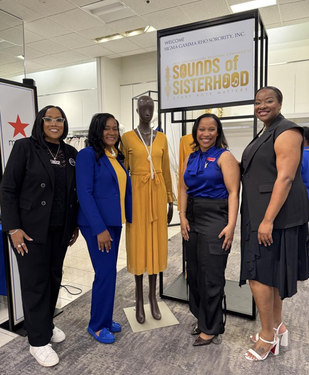 Five women standing next to a mannequin dressed in a yellow dress at the 'Sounds of Sisterhood' event by Sigma Gamma Rho Sorority, Inc., with a welcoming sign in the background and a clothing rack on the left.