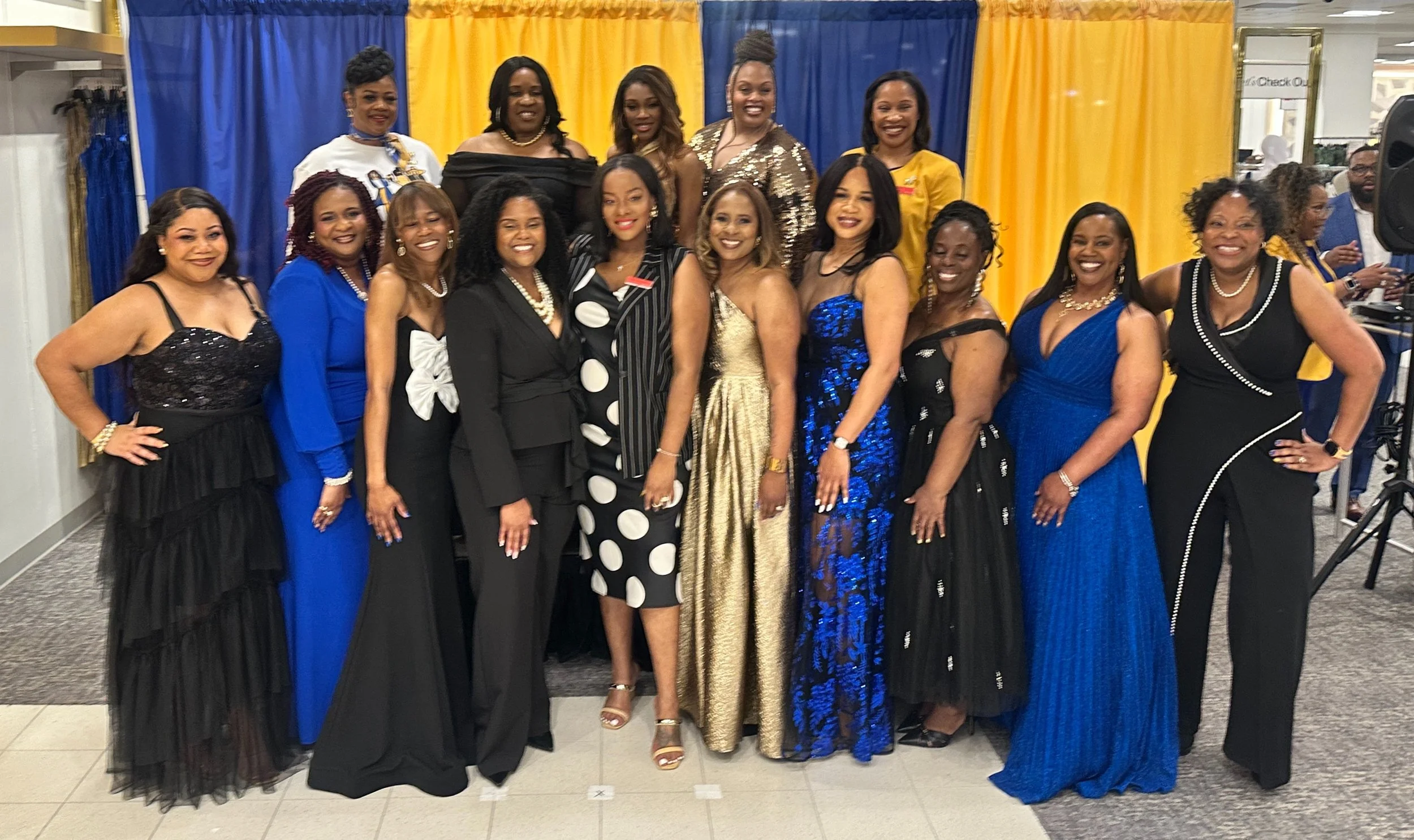 A group of women dressed in formal gowns and elegant attire posing together indoors in front of a blue and yellow decorated backdrop.