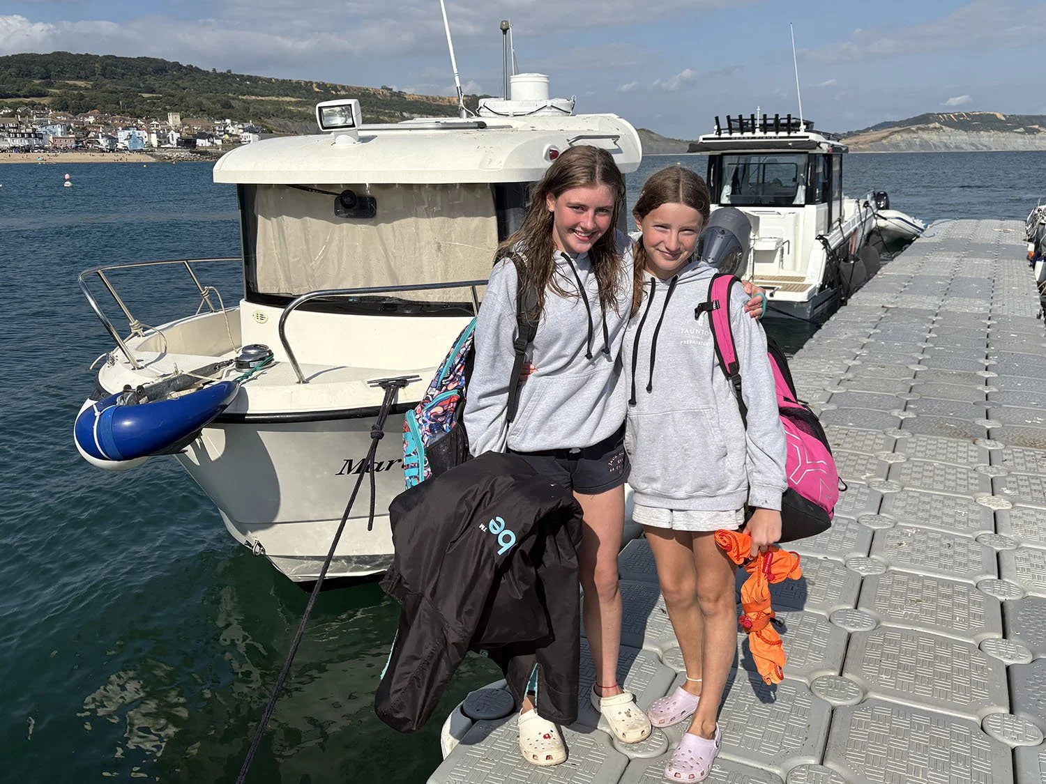 Two girls with backpacks and casual clothing standing on a dock by boats, smiling at the camera during daytime.