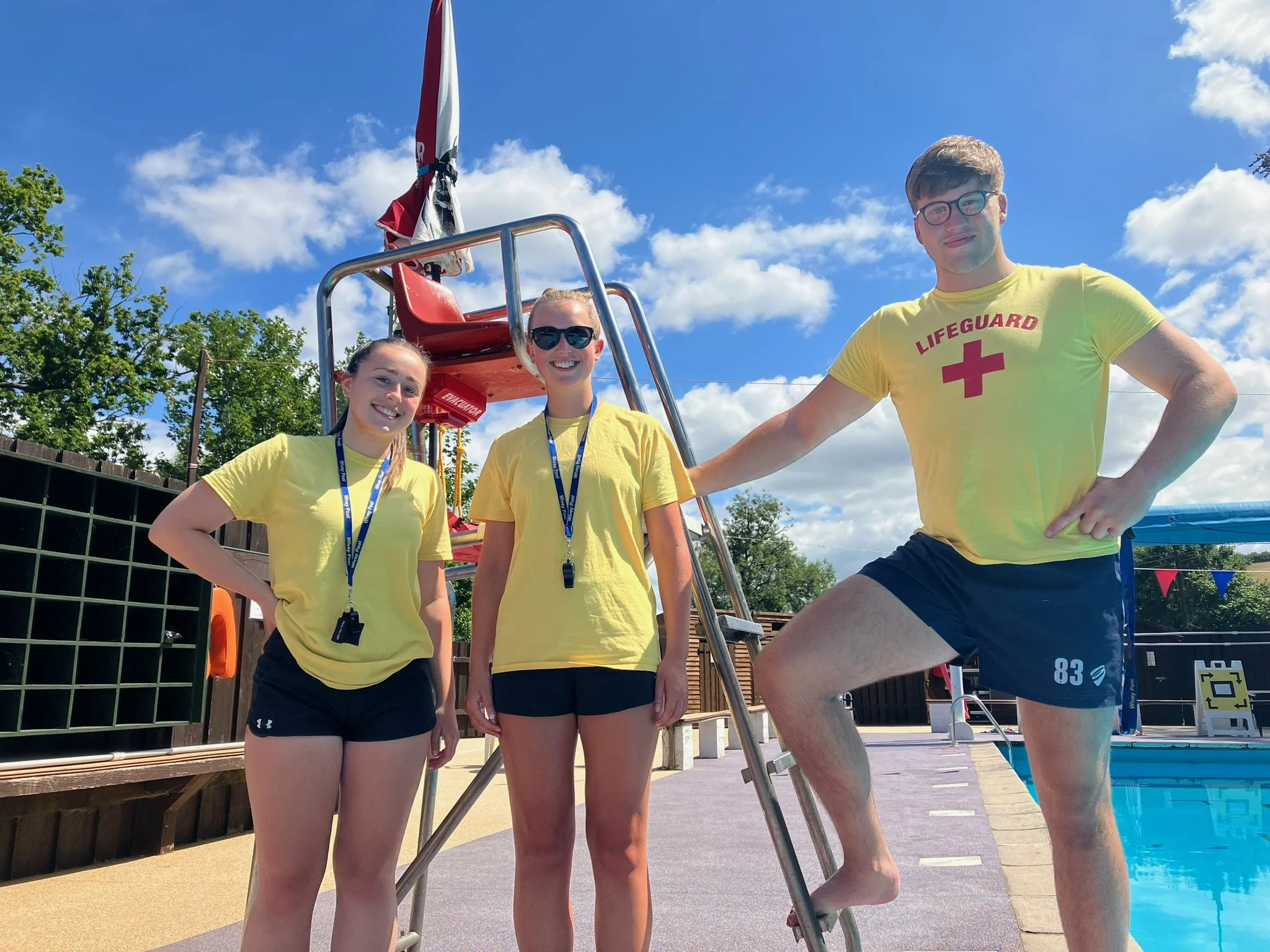 Three lifeguards standing near a swimming pool on a bright, sunny day. They are wearing yellow shirts and black shorts, with one of them wearing sunglasses. There is a red rescue chair and a banner in the background.