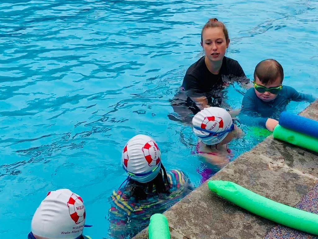 A young female swim instructor teaches children how to swim in a pool. The children are wearing swim caps and goggles, and are holding onto pool noodles near the pool's edge.