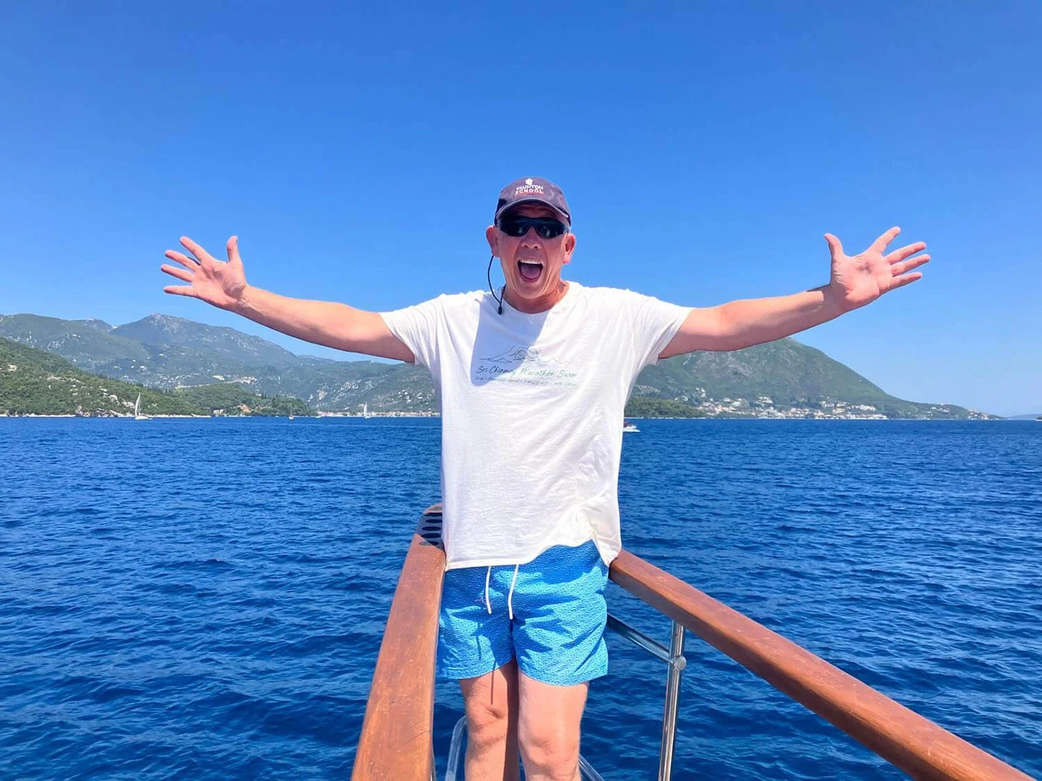 A man on a boat with arms outstretched, smiling, wearing a white t-shirt, blue shorts, a dark cap, and sunglasses, with a mountain landscape and blue water in the background.
