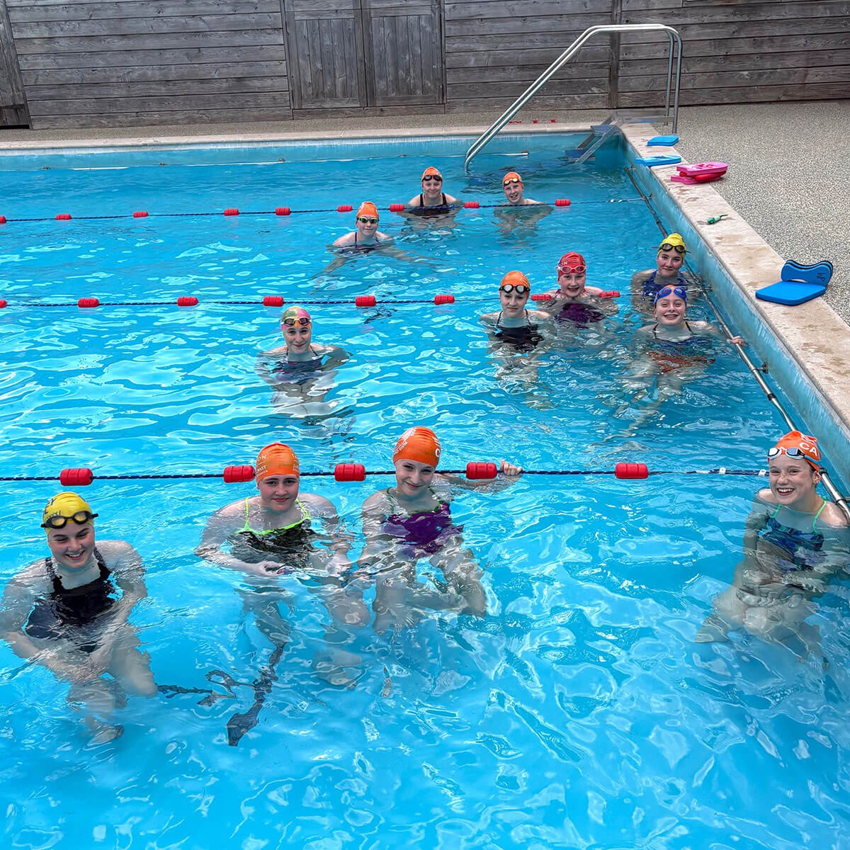 Group of young girls and women in a swimming pool wearing swim caps and goggles, participating in a swimming class or practice.