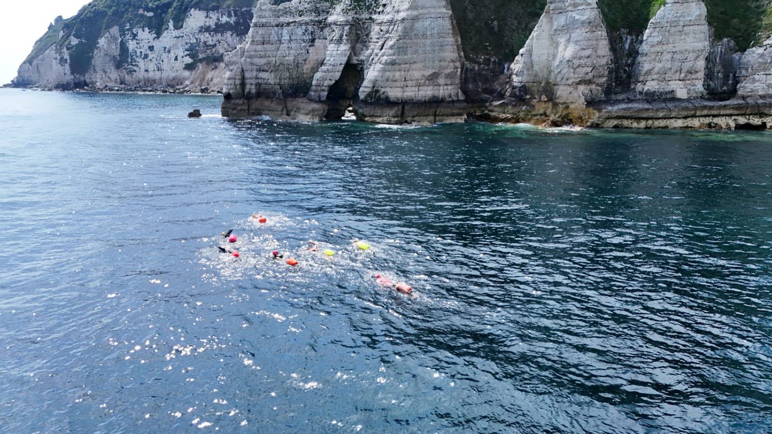 Seven swimmers with bright safety floats swim in the sea past cliffs at Beer in Devon, UK