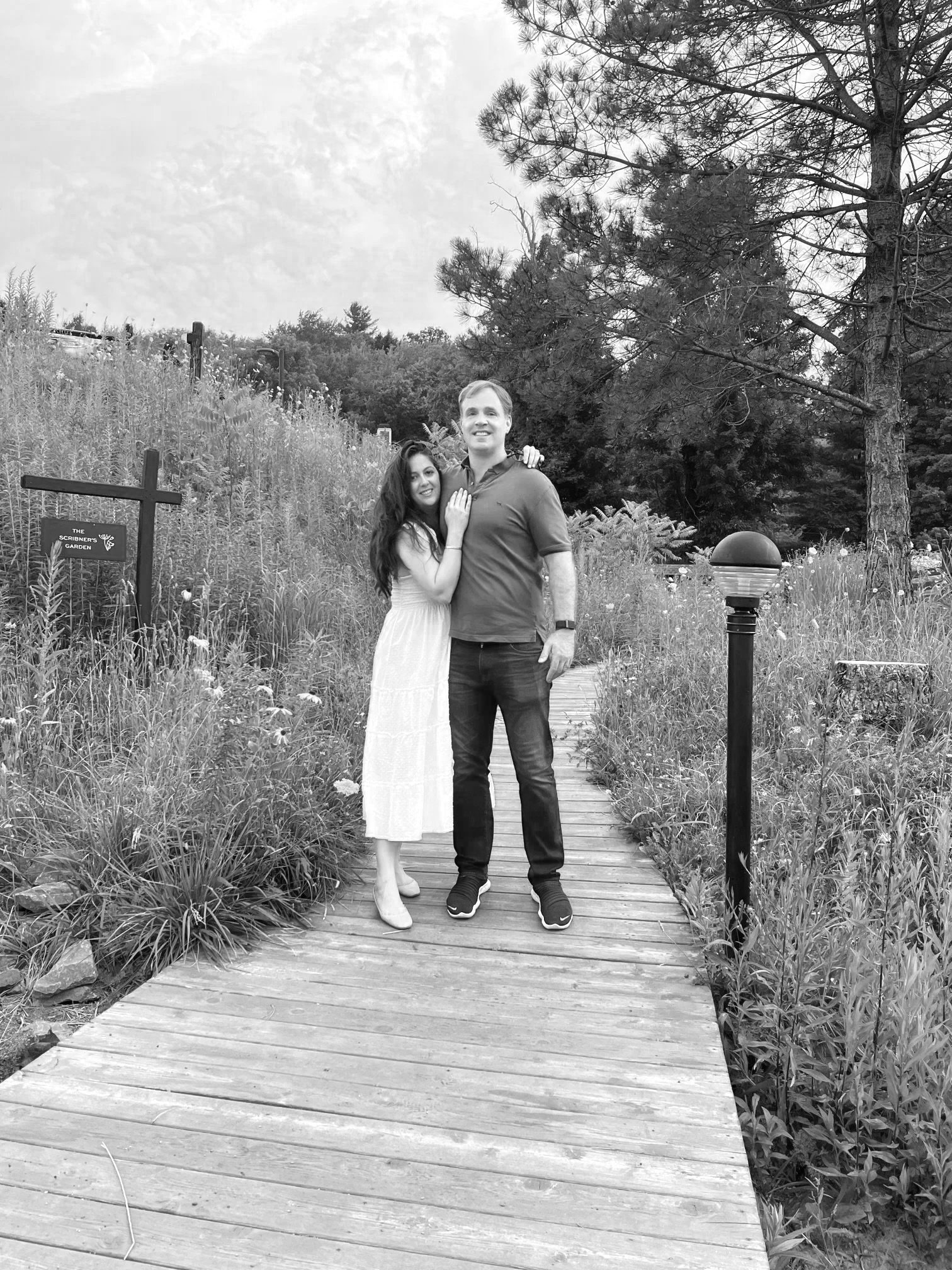 A couple standing on a wooden bridge in a garden, smiling at the camera, surrounded by tall grass and trees.