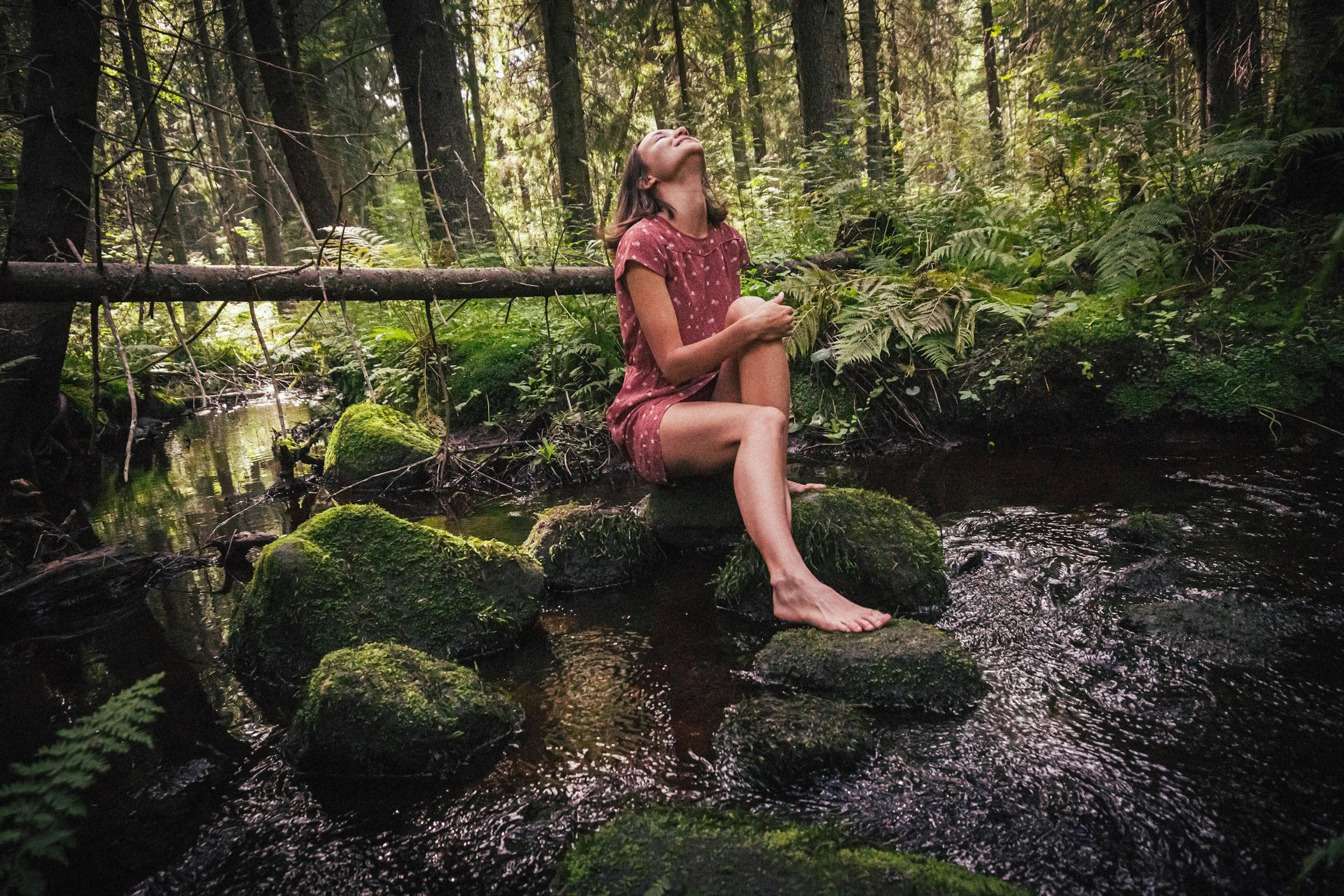 A woman sitting on rocks in a forest stream, with her eyes closed and head tilted back, wearing a red dress with white polka dots, surrounded by green foliage and tall trees.
