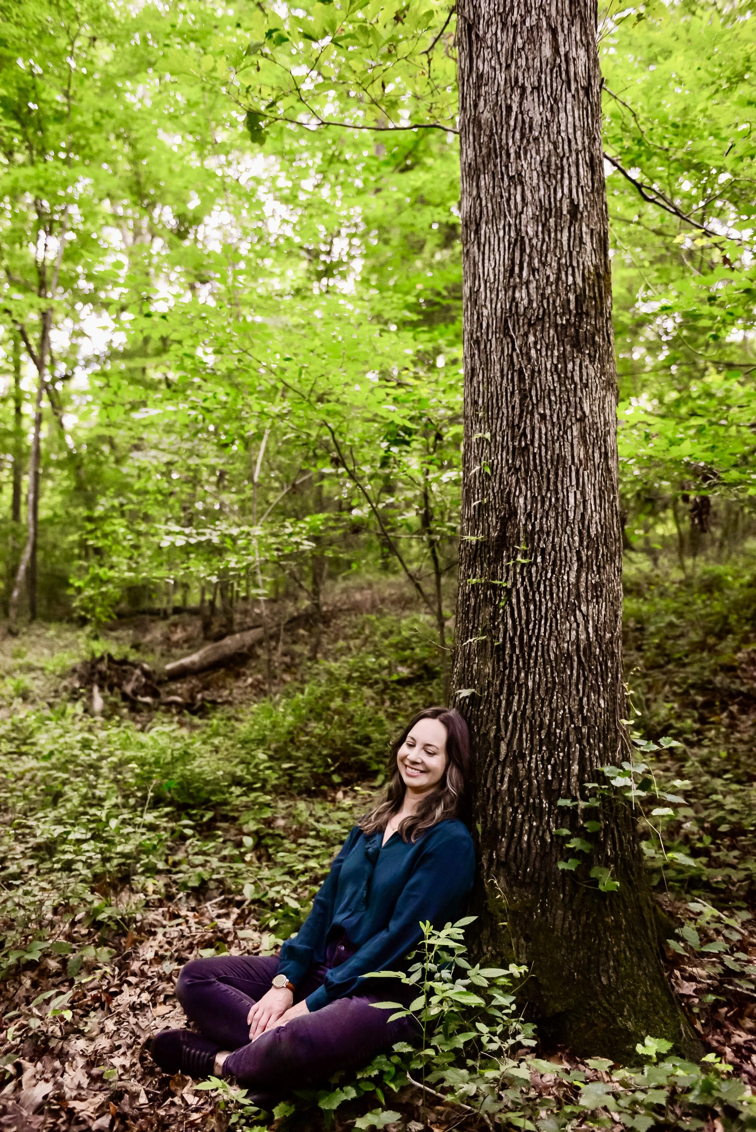 A woman with long brown hair, wearing a dark blue jacket and purple pants, sitting with her back against a tree trunk in a lush green forest. She has a smile on her face and her eyes are closed.