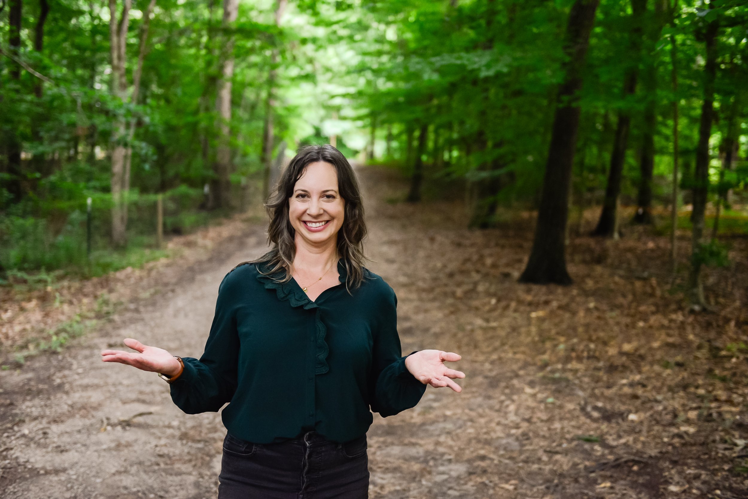 A woman with brown hair smiling and gesturing with her hands while standing on a dirt path in a green forest.
