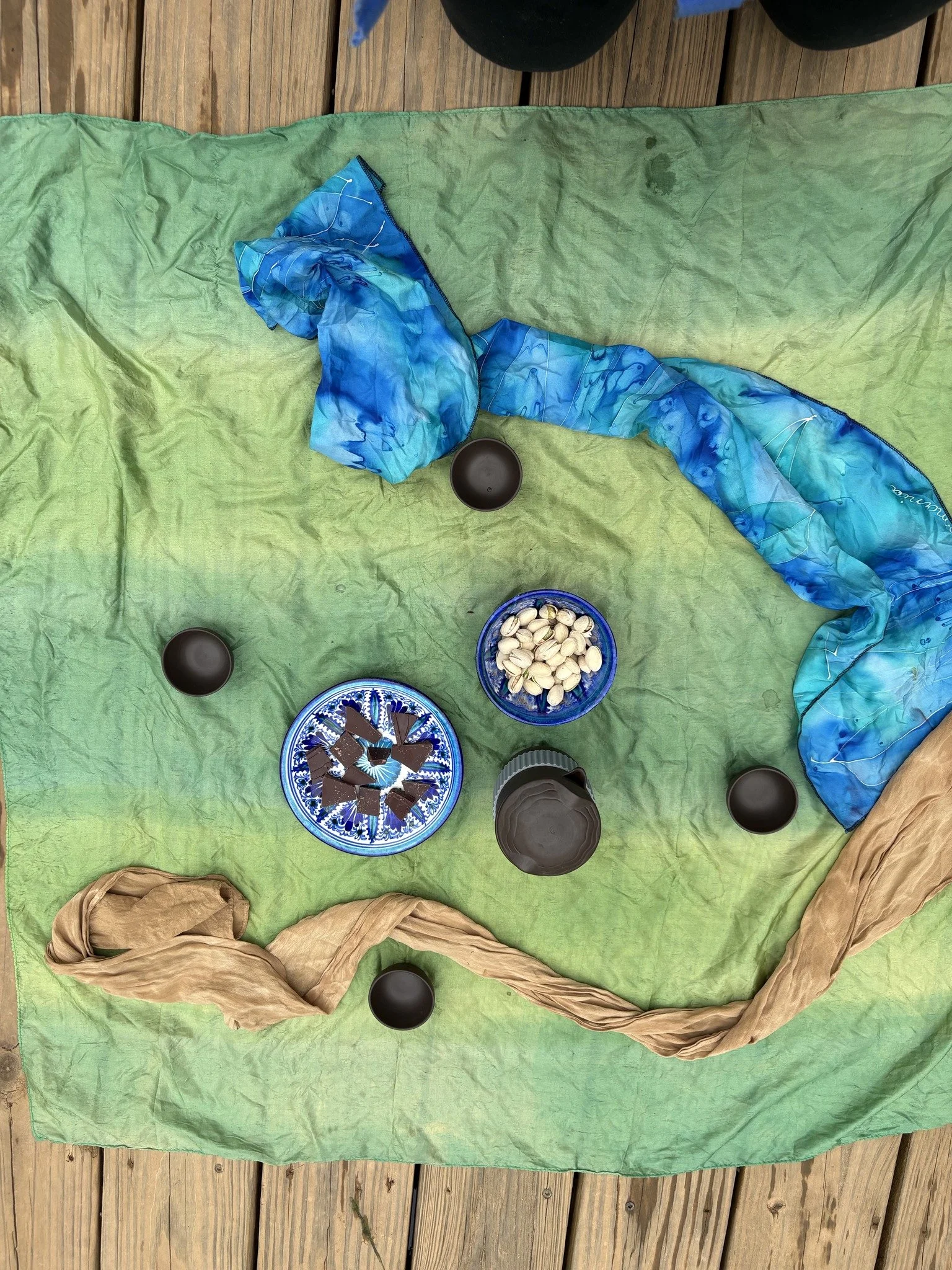 Colorful cloths and bowls with chocolate pieces and pistachios on a green cloth spread on a wooden deck.