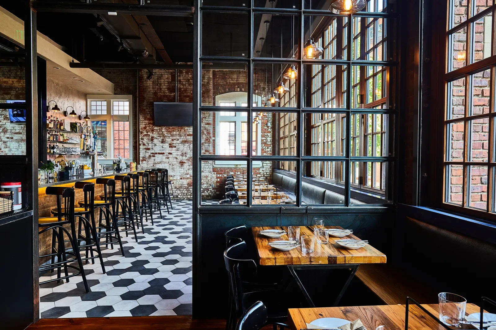 Interior of a modern restaurant with large windows, exposed brick walls, and a bar area with black barstools. The space has a mix of high tables with chairs and a cozy booth seating area. The decor features industrial style lighting and black-framed glass partitions.