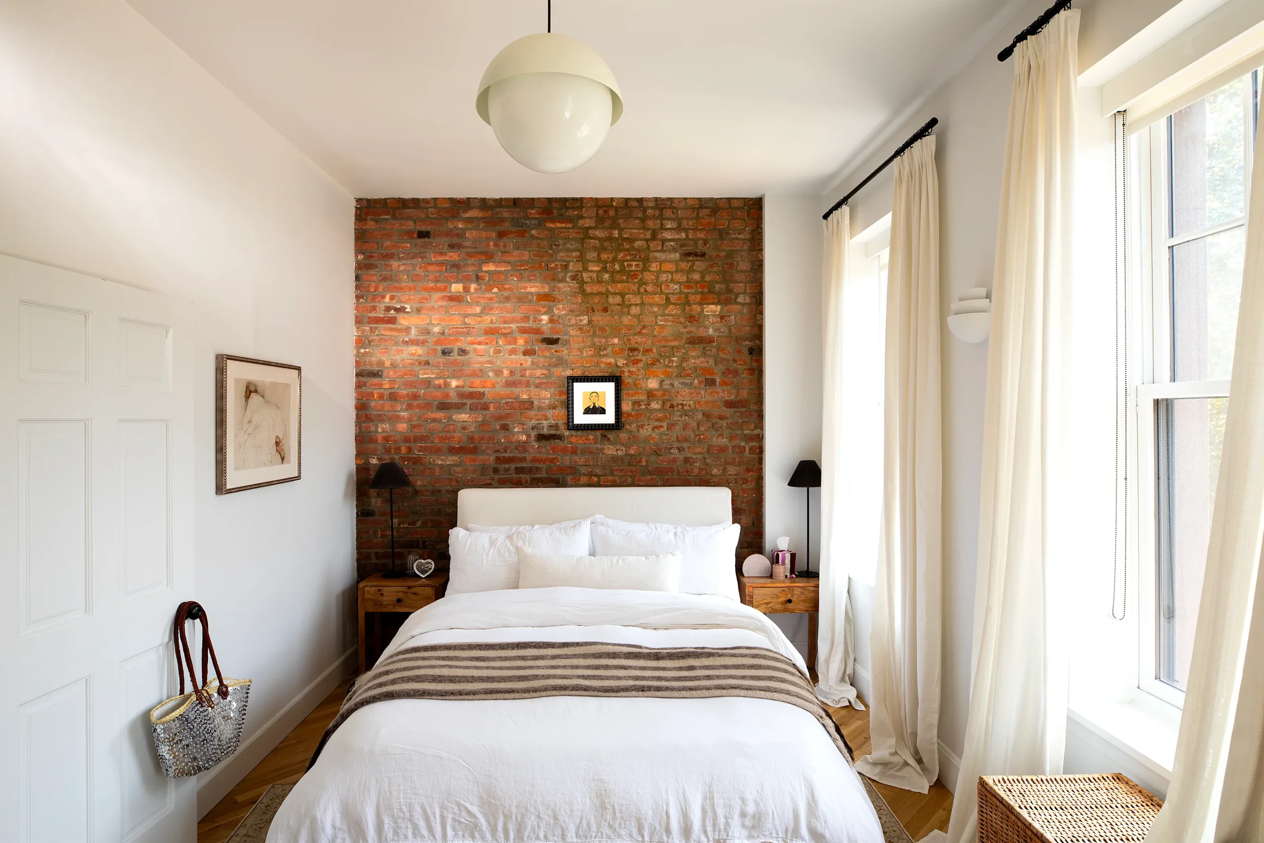 A bedroom with white walls and a bed with white bedding, a wooden headboard, and a white pillow. There are two small wooden nightstands on either side of the bed, each with a black lamp. An exposed red brick wall serves as the headboard area, with a small framed picture on it. Light streams in through large windows with cream curtains. There is a wicker basket near the window and a decorative bag hanging on a hook on the wall.
