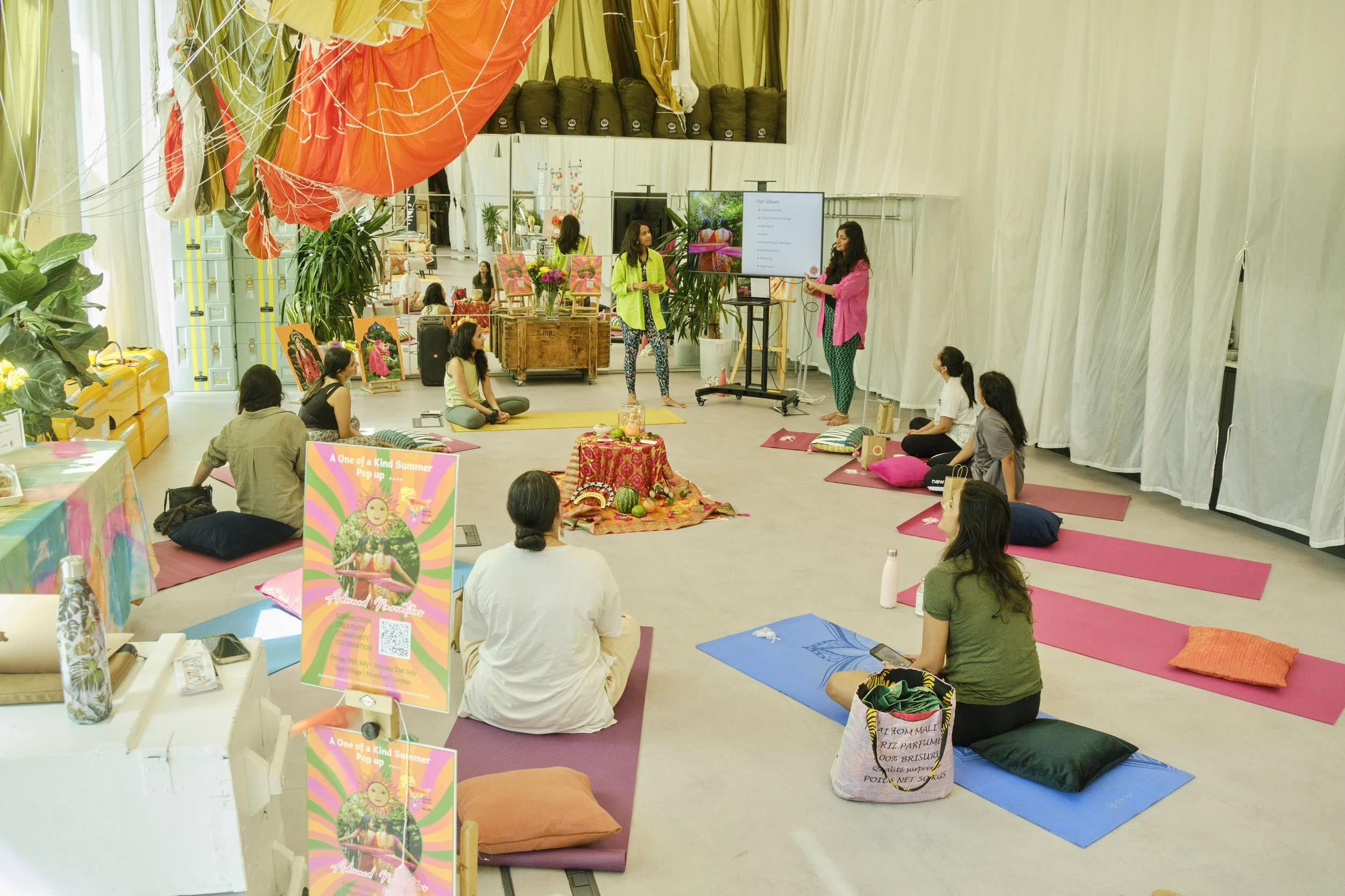 Group of people participating in a yoga or meditation class inside a bright, colorful space with decorations and artwork, some sitting on yoga mats and cushions while instructors lead the session.