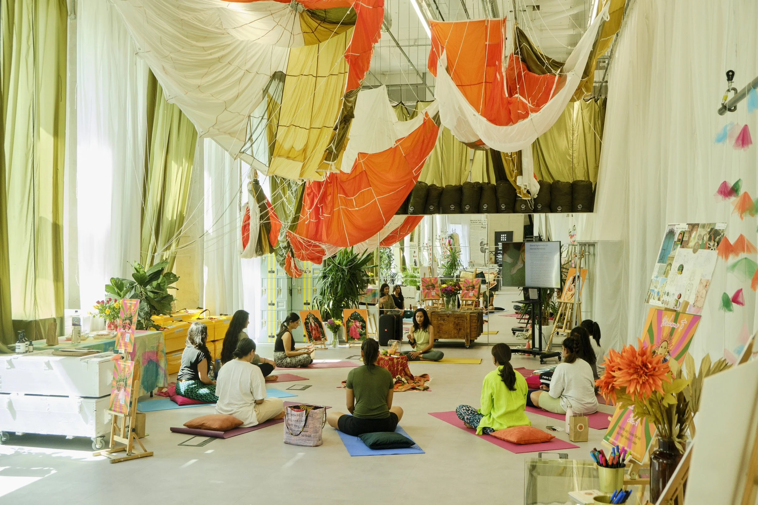 People seated on yoga mats participating in a meditation session inside a decorated indoor space with hanging fabric art and plants.