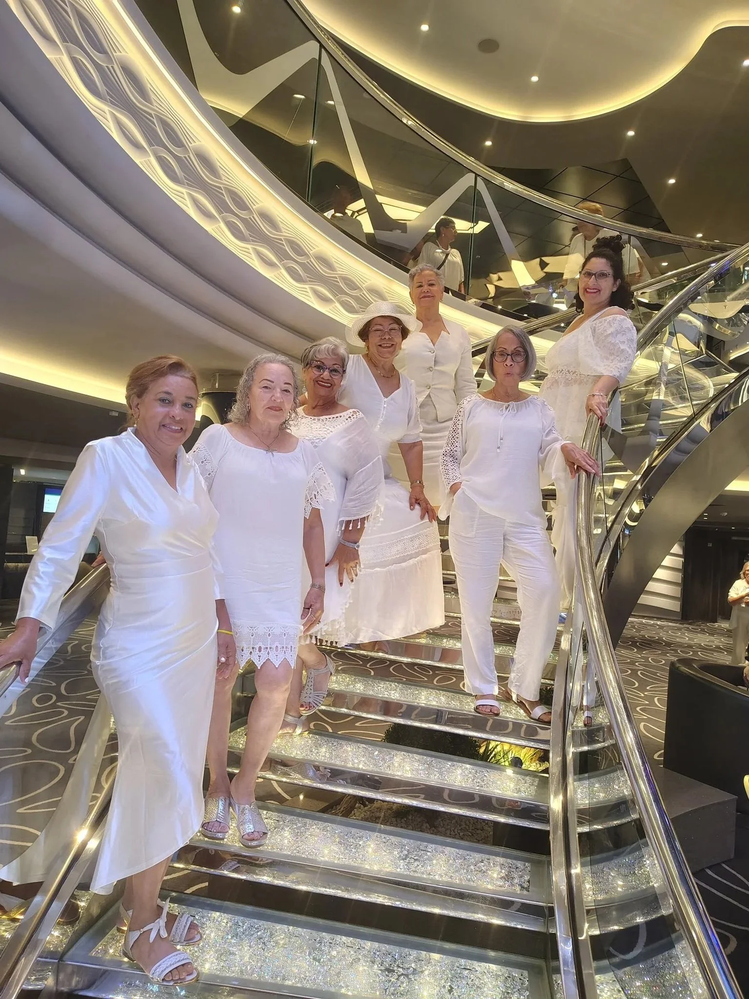 A group of 8 happy women dressed in white standing on the Swarovski Crystal Staircase inside MSC Grandiosa.